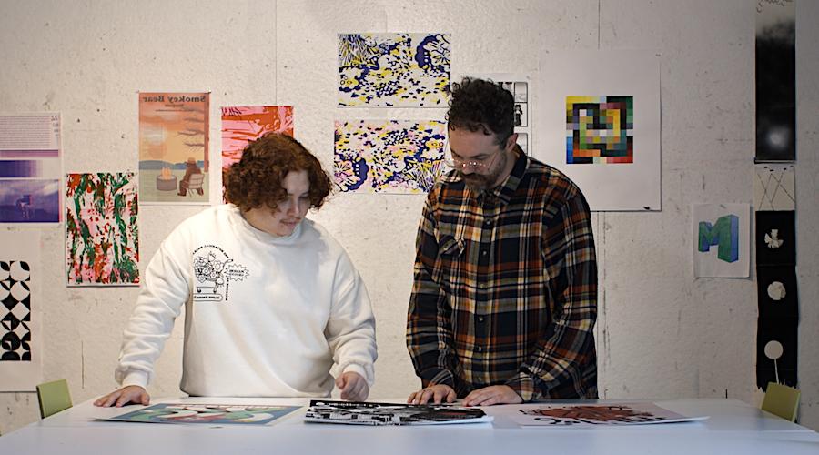 A student and professor look at prints at a large table. There is digital artwork hanging on the wall behind them.